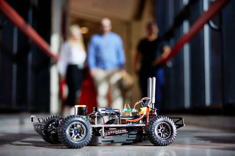 closeup of small autonomous racing car with three figures blurred in background